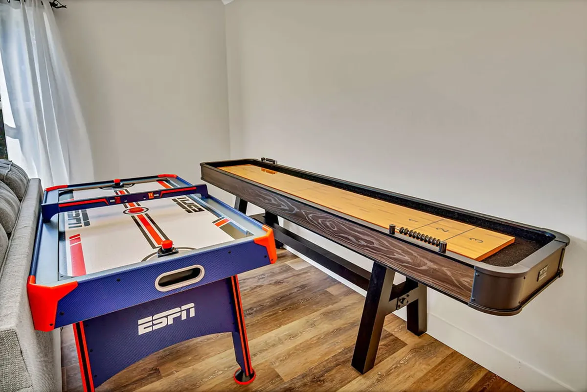 The photo highlights an air hockey table and shuffleboard table alongside a comfy sofa. The setup evokes a welcoming, playful atmosphere, perfect for hosting tournaments or casual family fun, reinforcing the room’s all-ages appeal.