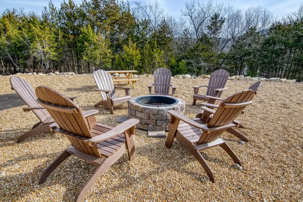 An outdoor fire pit surrounded by Adirondack chairs and encircled by trees, creating a cozy and inviting atmosphere for socializing, storytelling, or stargazing under the open sky.