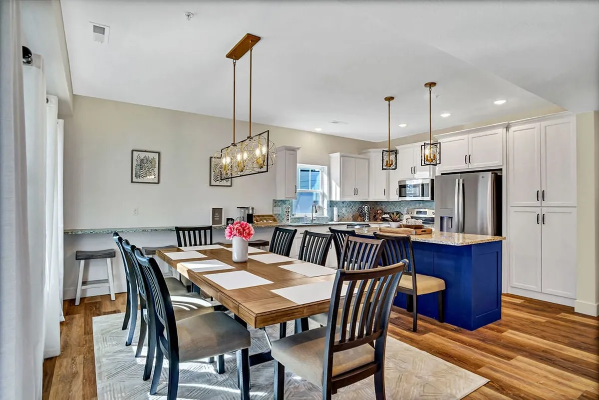 A stylish dining area with an elegant table set for eight, adjacent to a spacious kitchen island with bar seating. Modern lighting fixtures and an abundance of natural light create an inviting atmosphere for group meals and gatherings.