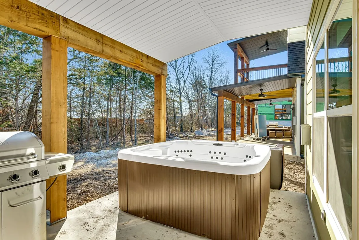 An exterior shot featuring a bubbling hot tub next to a grill and outdoor lounge seating under rustic wooden beams. The space overlooks nature and invites guests to unwind outdoors, especially at sunset.