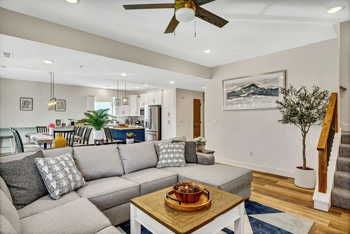 A cohesive open-plan interior showing the sectional sofa, dining area, kitchen island, wall art, and an indoor plant, all illuminated by natural light from large windows. The layout demonstrates the seamless connection between living, dining, and kitchen spaces.