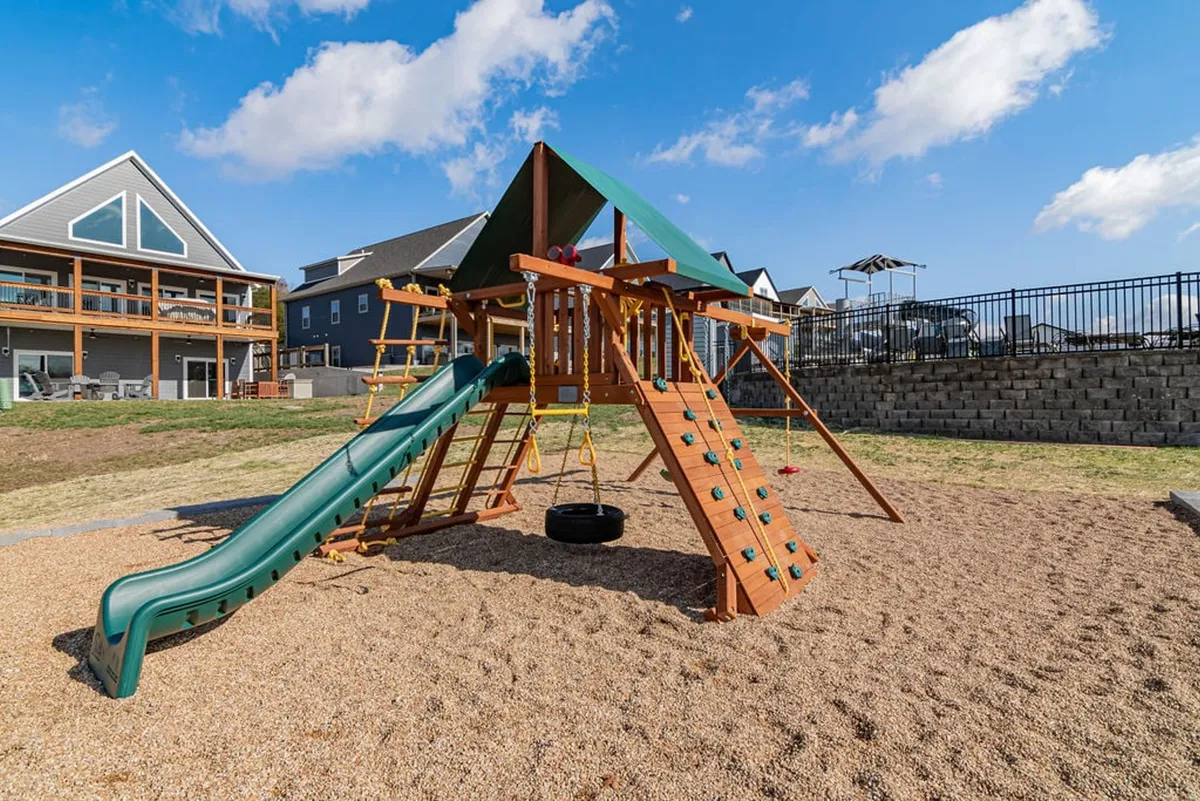 An exterior shot of the playground area, highlighting play structures including a slide, swing set, and sandbox. Children are captured in the midst of joyful play, while families gather and socialize nearby, all surrounded by a lush green setting.