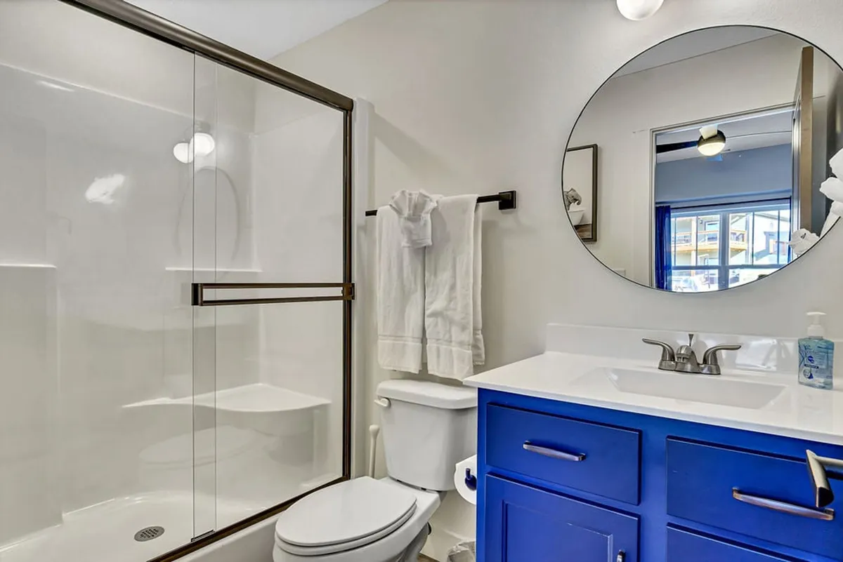 A modern en-suite bathroom featuring a blue vanity cabinet, round mirror, and a sleek glass-enclosed shower. The space is clean and inviting, suggesting a spa-like atmosphere for guests.