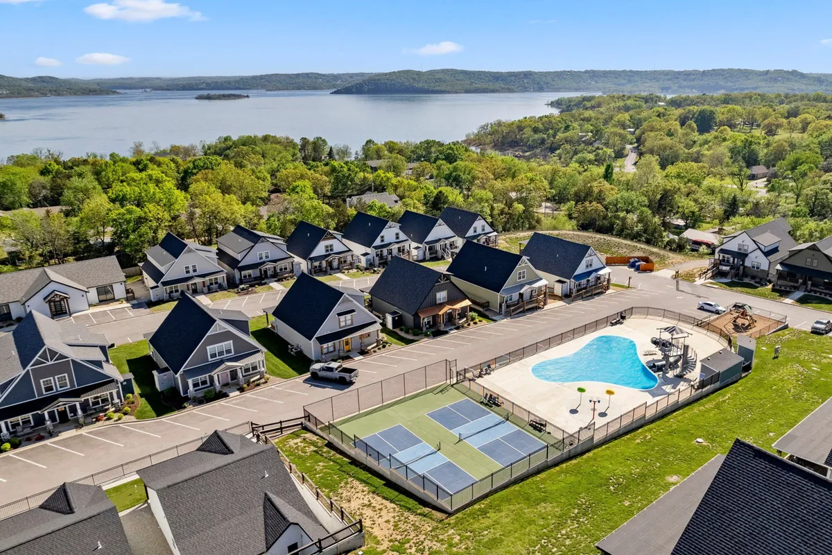 A lively exterior scene featuring a community pool, tennis courts, surrounding homes, and a scenic lake view. The area is set up for group gatherings, swimming, and friendly matches, encapsulating the active, communal amenities available nearby.