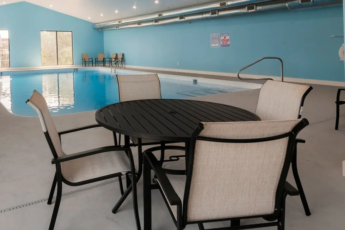 A bright and comfortable indoor swimming pool area, featuring a table and chairs for relaxation, large windows letting in natural light, and an inviting swimming space for year-round enjoyment.