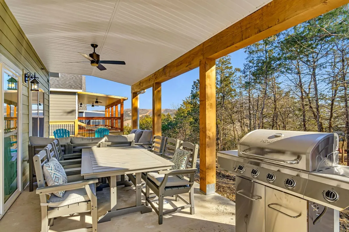 A spacious deck just outside the main living area, featuring an outdoor dining table, gas grill, and comfortable seating, with a backdrop of lush trees. The space is ideal for alfresco meals and gatherings under the sky.