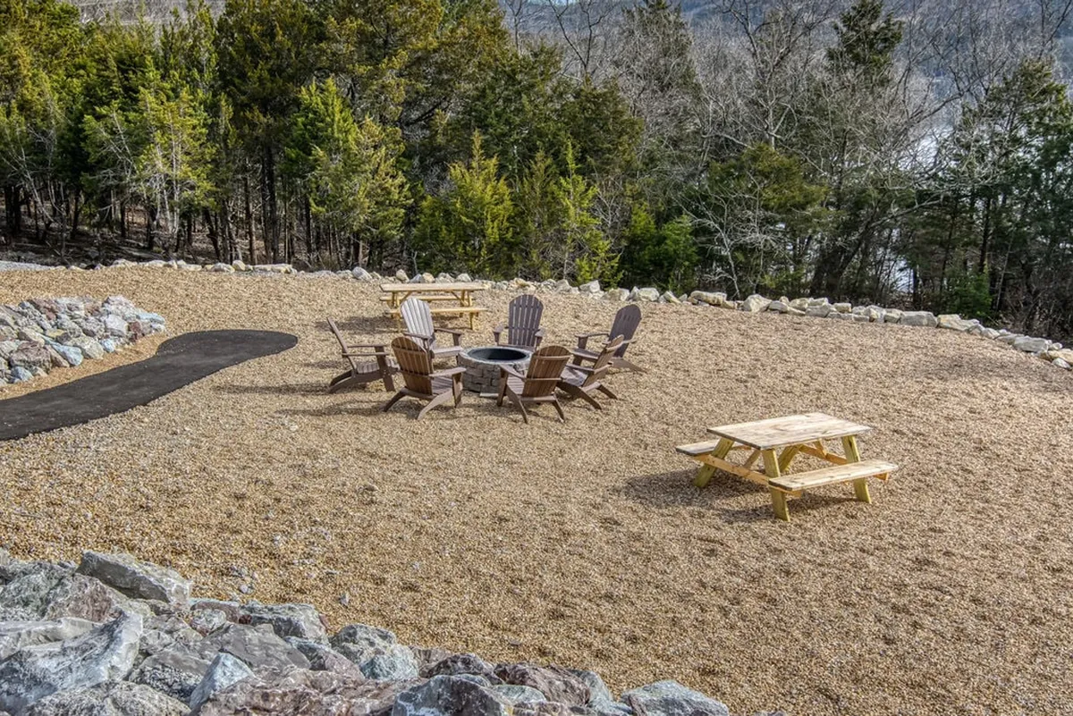 A cozy fire pit area set among wooden chairs and a picnic table, with a winding pathway and greenery nearby. The scene invites relaxed evenings spent gathering and making memories around the fire.
