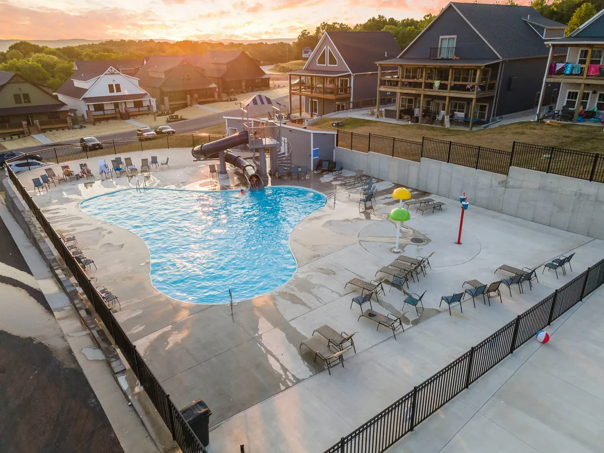 A vibrant exterior view of the outdoor swimming pool area, featuring a lively water slide, colorful water features, and plenty of lounge chairs. The setting is bustling with activity, with kids and families swimming, playing, and socializing, all under the glow of a beautiful sunset.