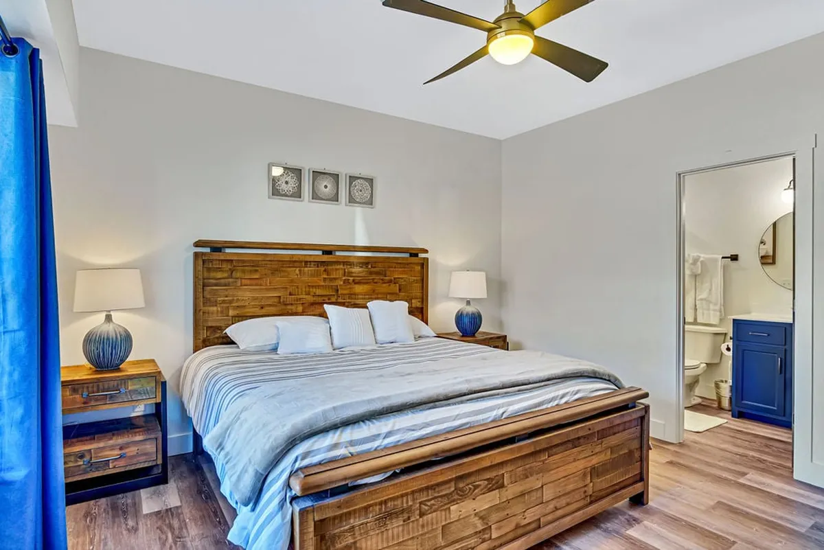 A serene king bedroom featuring a wooden bed frame, blue curtains, decorative wall art, and nightstands with lamps. The setting is warmly lit, with natural light filtering through the window, creating a peaceful and cozy ambiance ideal for relaxation.