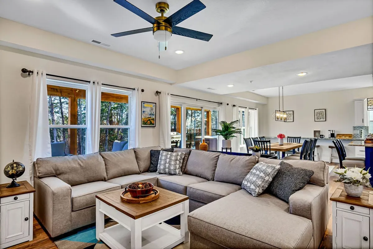 The image showcases a spacious, sunlit living and dining area featuring a plush sectional sofa, a large wooden dining table perfect for gatherings, and expansive windows that fill the space with natural light. The setting exudes warmth and comfort, inviting guests to relax, socialize, and make lasting memories together.